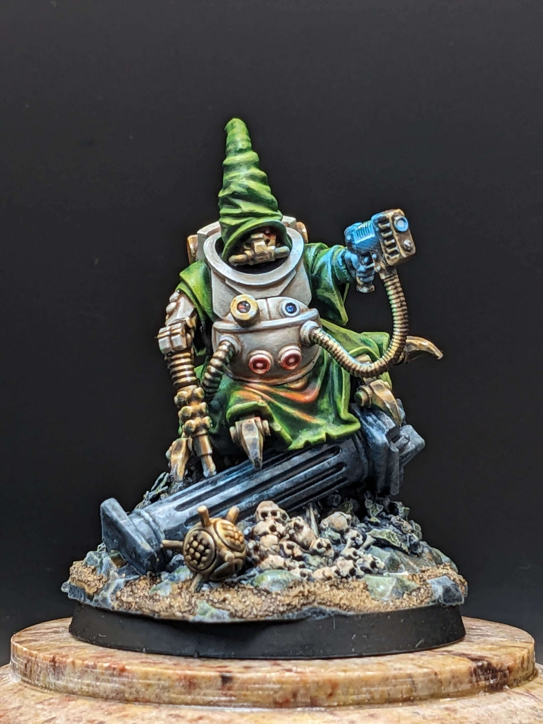 Painted competition miniature of a character in a green robe and pointed hat, seated on rubble with a futuristic gadget.